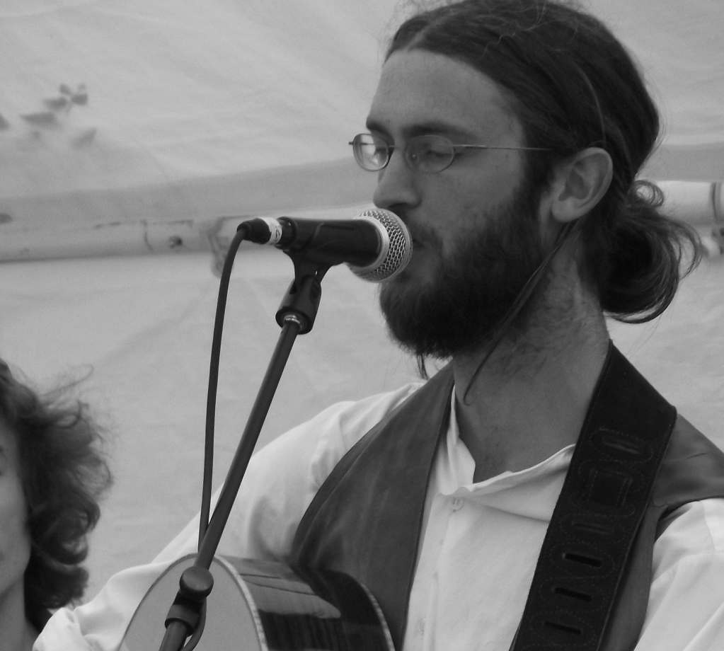 A man with dark hair in a ponytail, beard and glasses singing into a microphone while playing an acoustic guitar, with a blurred figure in the background.