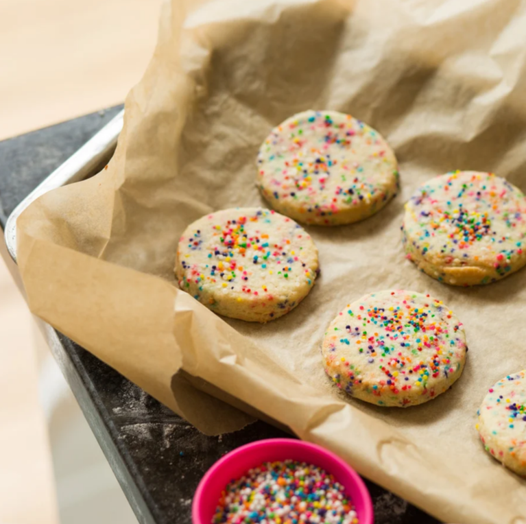 Image shows four baked biscuits on brown parchment paper on a baking tray. Each biscuit has multi-coloured sprinkles on and there is a pot of sprinkles to the left of the baking tray.