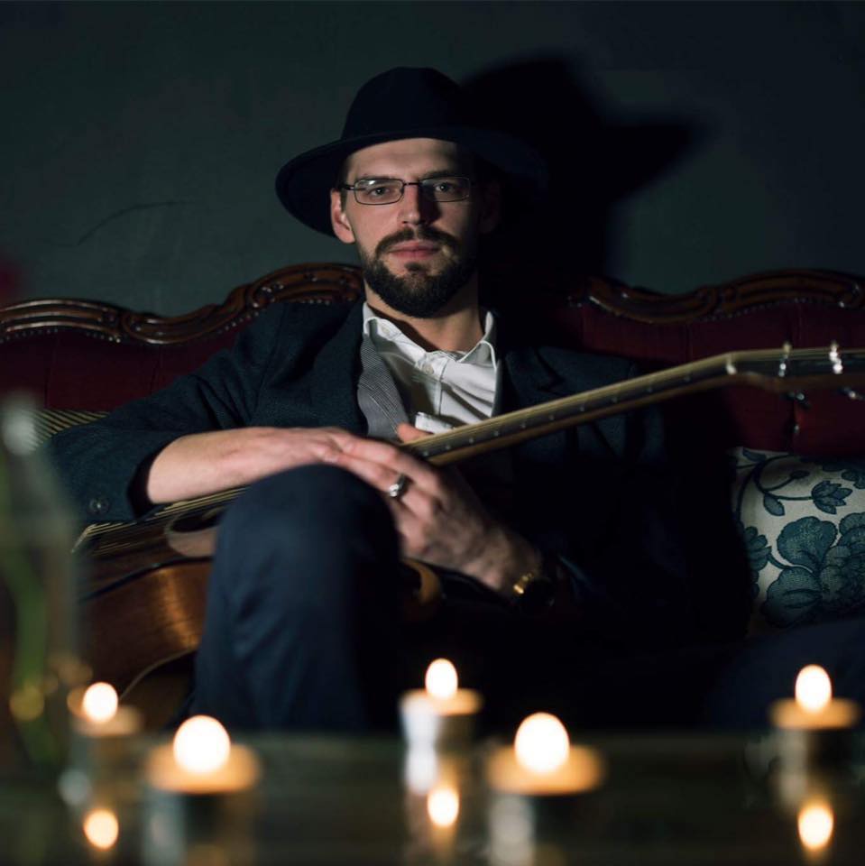 A man with a beard, glasses, and a black hat sits on a vintage couch holding a guitar, surrounded by softly glowing tealight candles.
