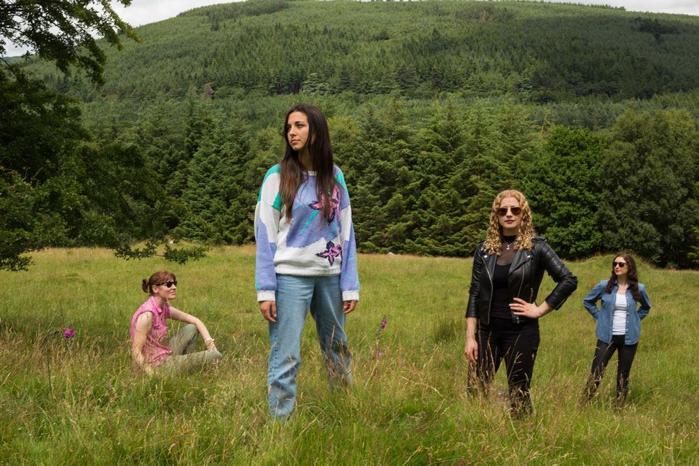 Four women standing and sitting in a grassy field surrounded by trees and a hilly landscape in the background, dressed in casual attire.
