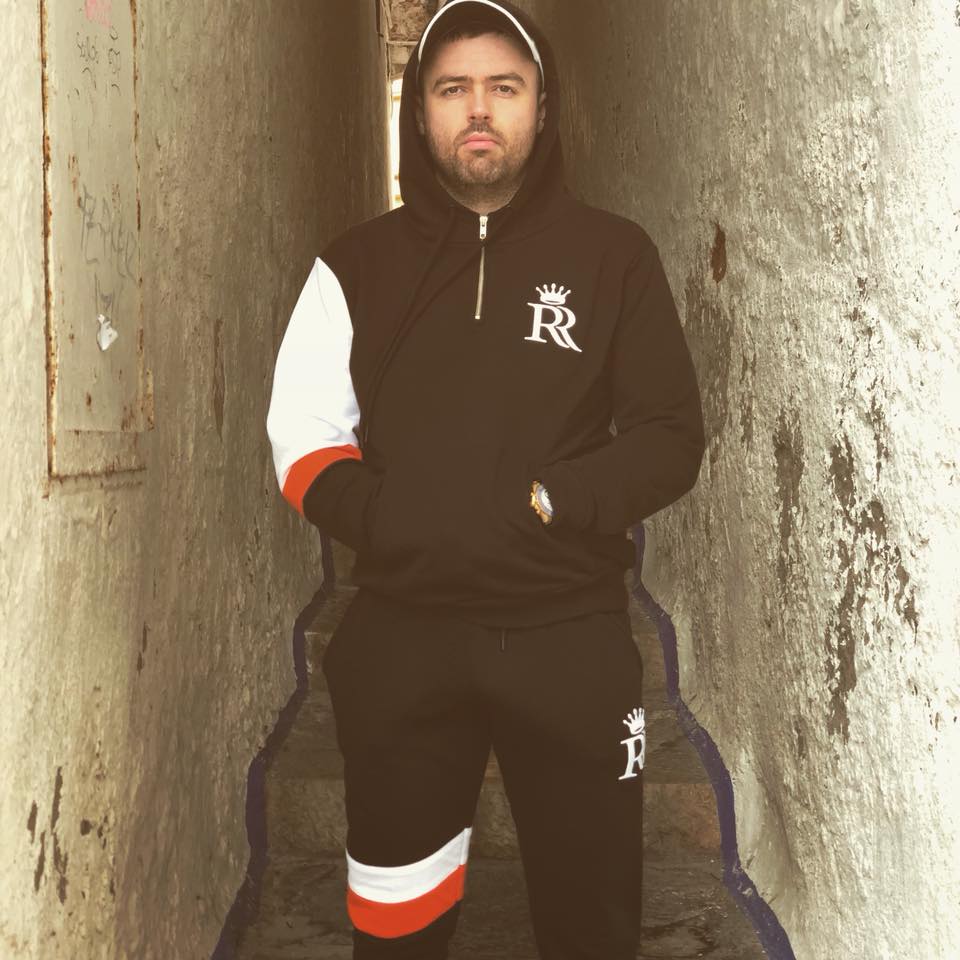 A man with dark hair and a beard standing in a narrow alleyway, wearing a black zip-up hoodie and matching sweatpants with colourful stripes. He has a serious expression and is holding his hands in his pockets.