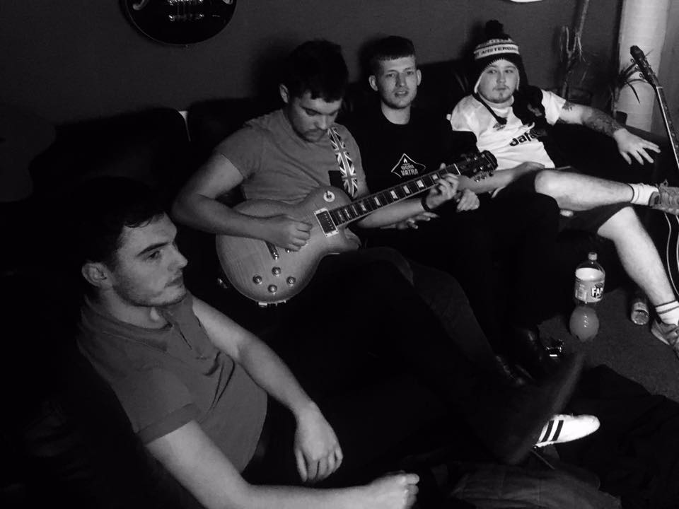 A black and white photo of four young men sitting on a sofa in a dimly lit room; one is playing an electric guitar, while the others are relaxed and watching.