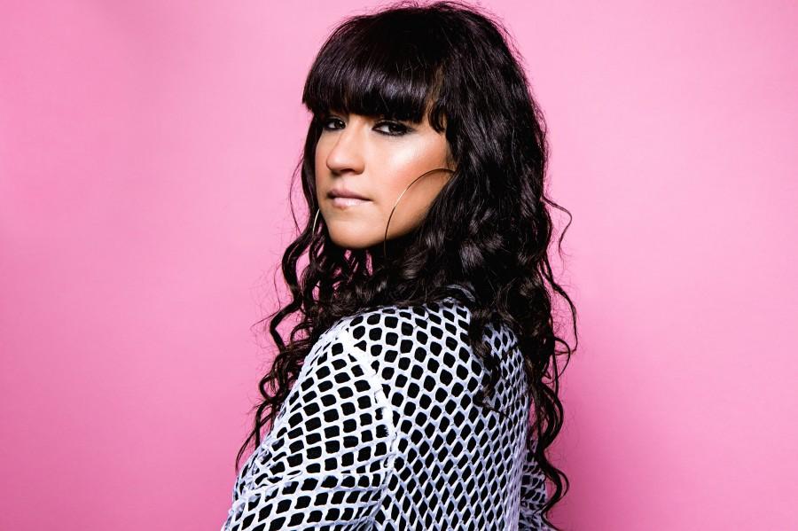 A woman with long, curly hair poses against a pink background, wearing a patterned top and large hoop earrings.