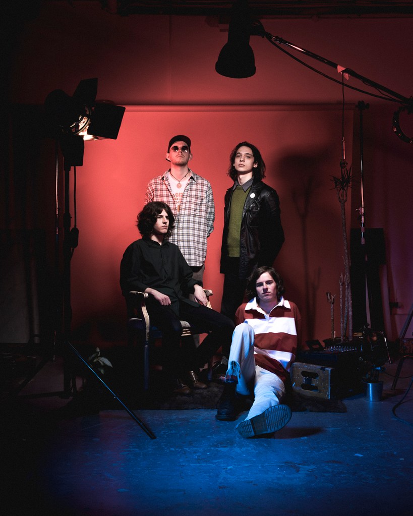 A group of four young men posing in a studio setting with dramatic lighting. The background is a gradient of red and blue, and they are dressed in fashionable, varied outfits, standing and sitting on props.