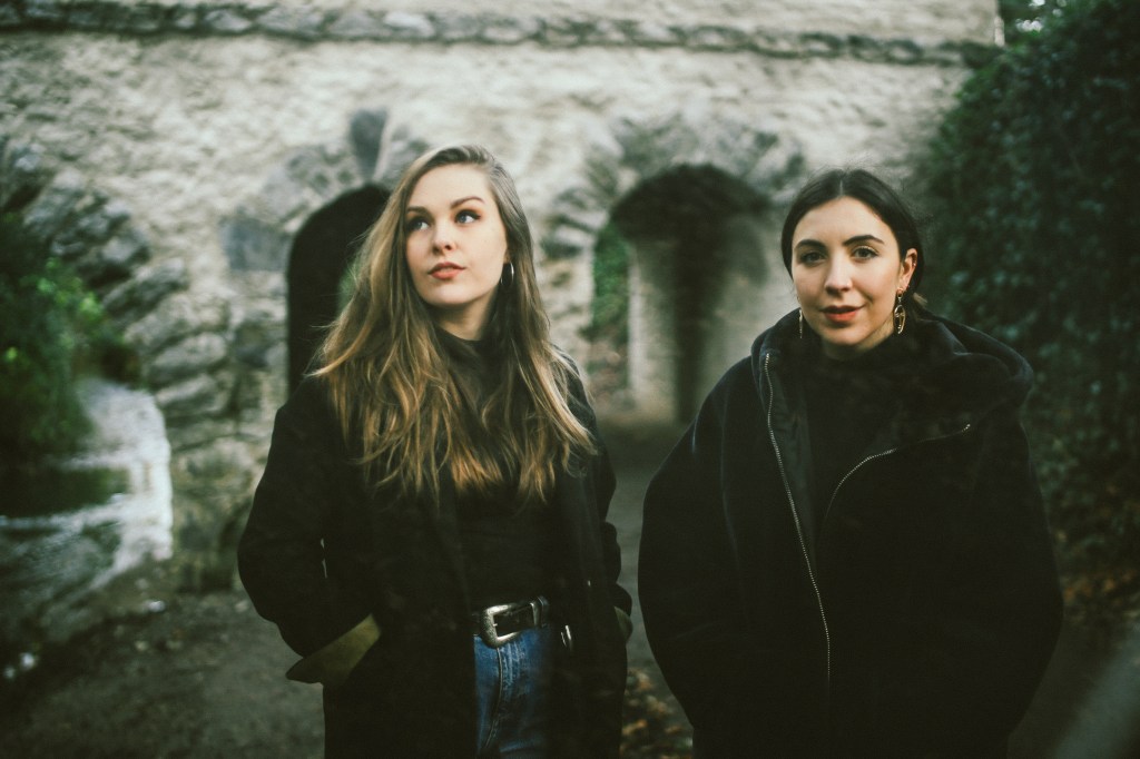 Two young women standing in front of an archway made of stone, surrounded by greenery, with a moody atmosphere.