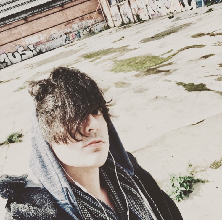 A man with long dark hair wearing a hoodie and earphones stands in an urban setting with graffiti in the background.