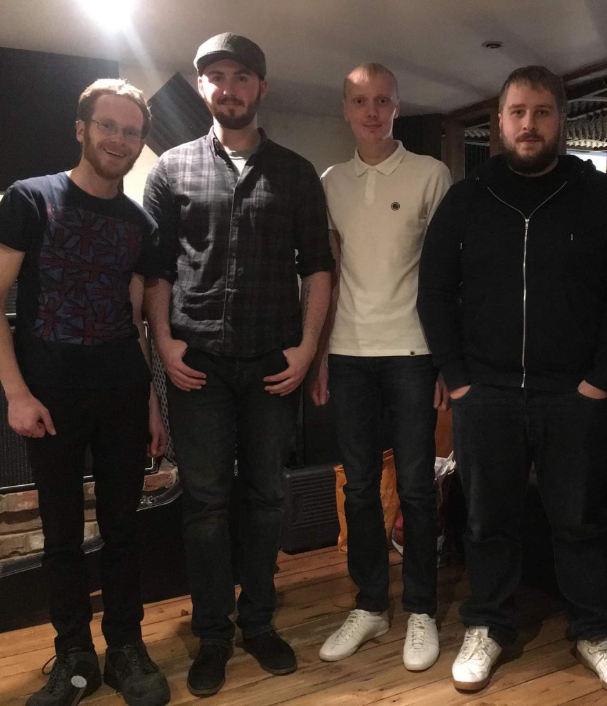 Four men standing next to each other in a casual setting, wearing a variety of shirts and jeans. The background features wooden flooring and partially visible furniture.