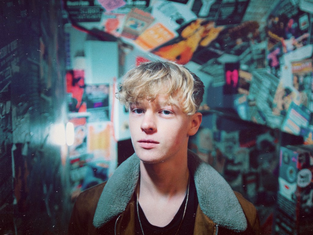 Portrait of a young man with tousled blond hair, wearing a brown jacket with a fur collar, standing in a narrow corridor filled with colourful posters and flyers on the walls.