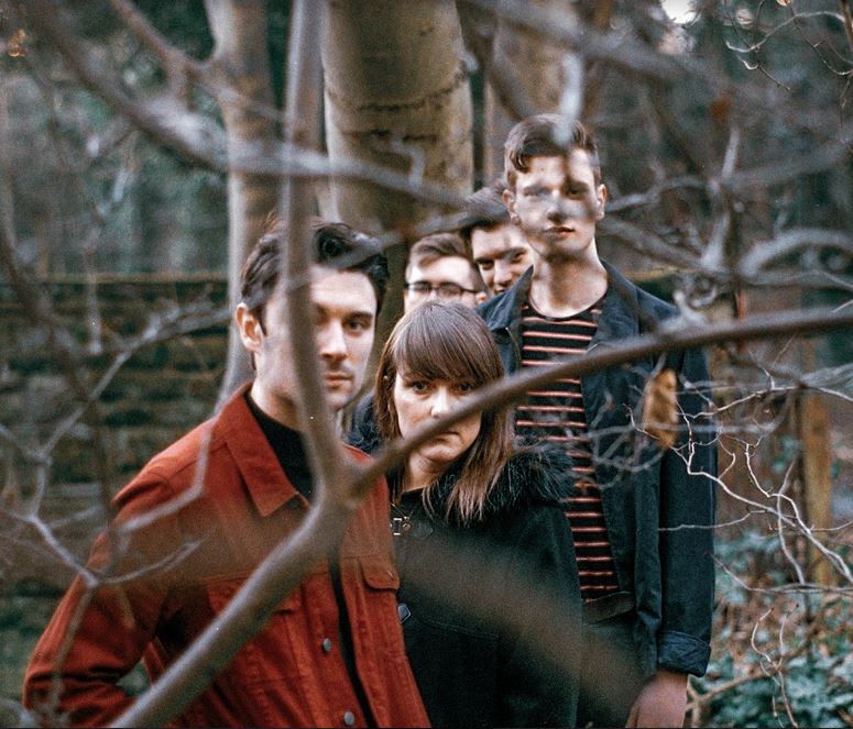 A group of five individuals standing closely together in a wooded area, partially obscured by branches. The foreground features a person in a red jacket, with others in dark clothing in the background.