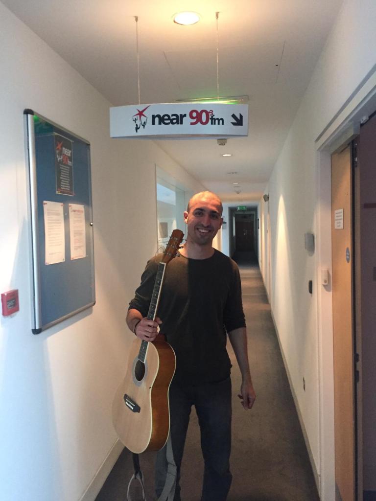 A man holding an acoustic guitar smiles while walking down a hallway with a sign for 'near 90fm' overhead.