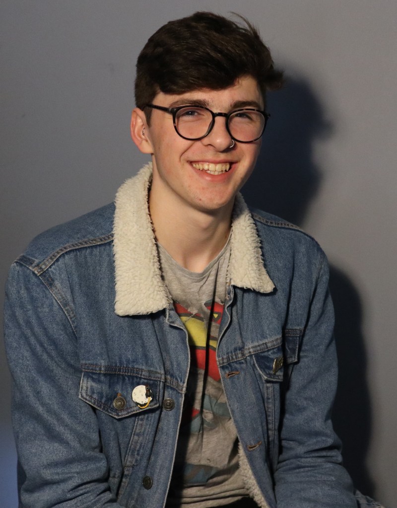 A young man with short dark hair, glasses and a friendly smile, wearing a denim jacket with a fur collar and a graphic t-shirt, sitting against a grey background.