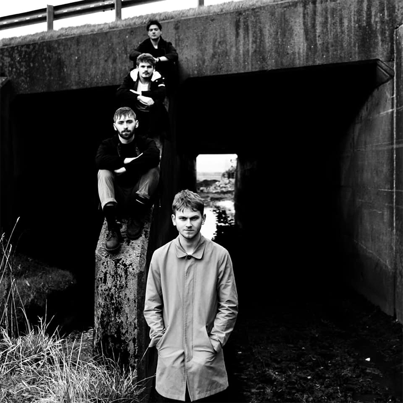 A black and white photo of four young men posing near a concrete structure, with one person standing in front and three seated or leaning behind them, framed by an opening in the structure.
