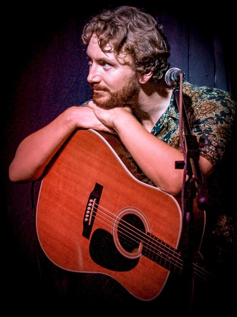 A musician with curly hair and a beard, resting his chin on his hands while holding an acoustic guitar, in a dimly lit setting.
