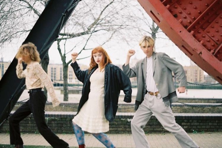 Three young adults striking dynamic poses outdoors near a large red structure, with trees and a waterfront in the background.