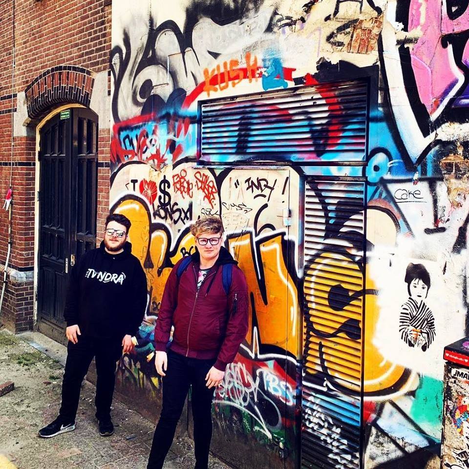 Two young men wearing black glasses, standing in front of a vibrant, graffiti-covered wall with various colourful tags and designs. The man on the left has short dark hair and wears a black hoodie top with the word 'TVNDRA' in white text, black trousers and black trainers. The man on the right has short light brown hair and wears a maroon zipped up jacket and dark trousers.