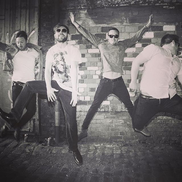 A black and white photo of four men jumping in their air against a brick wall, showcasing a dynamic and playful pose, with varying outfits and expressions.