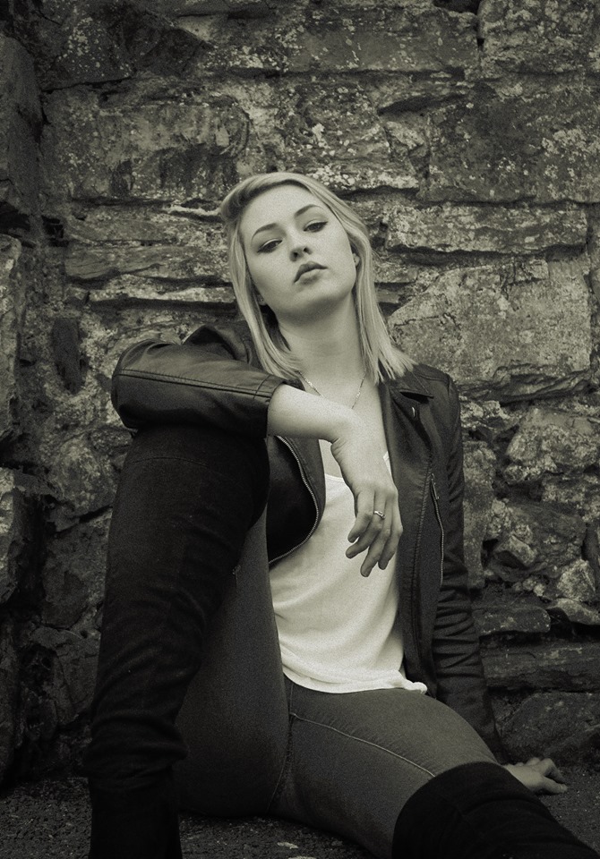 A black and white photo of a young woman with long blonde hair sitting against a stone wall, wearing a black leather jacket, white top and dark jeans, with a contemplative expression.