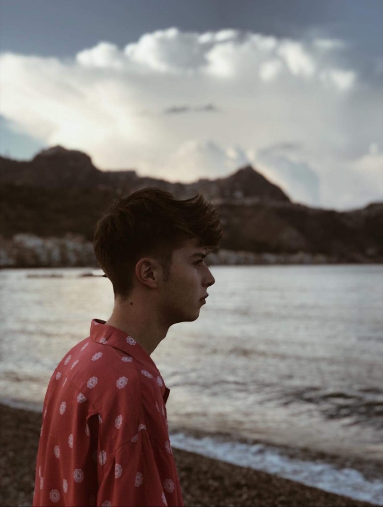 A young man with short dark hair in a pink patterned shirt standing by the water's edge, looking towards the sea with mountains and clouds in the background.