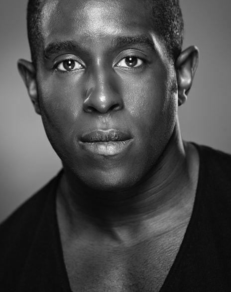 A close-up black and white portrait of a man with short hair, strong facial features, and a neutral expression, wearing a black v-neck shirt.