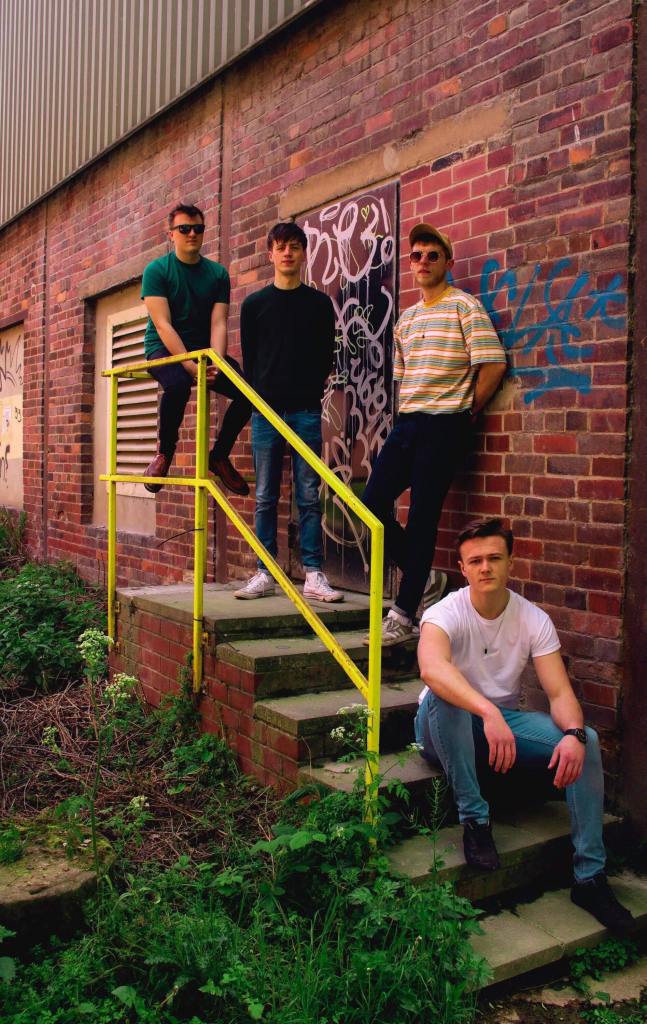 A group of four young men posing on a staircase with a yellow handrail in front of a brick wall covered with graffiti. Three are standing on the steps, while one sits at the bottom.