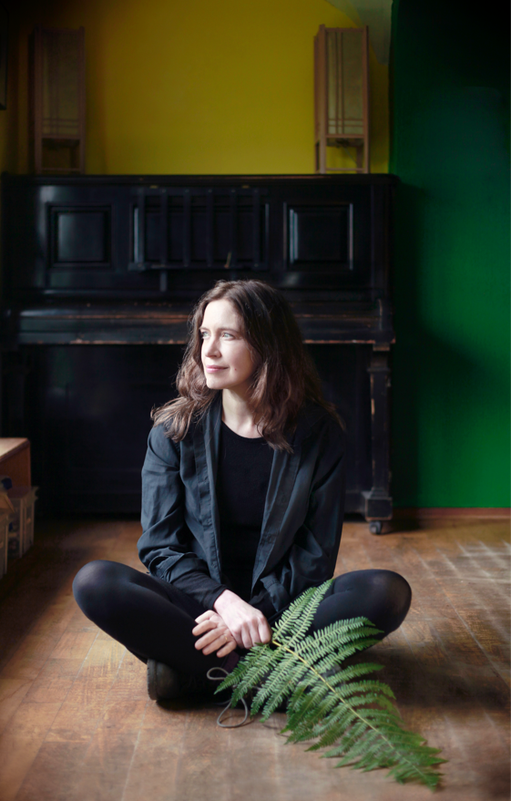 A woman with long wavy dark hair wearing a black outfit sits cross-legged on a wooden floor, holding a large fern. Behind her is a two-tone wall with yellow and green colours, and a dark piano in the background.