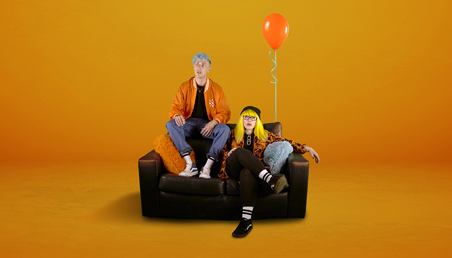 A man and woman sitting on a black sofa against a bright orange background. The man has blue hair and wears an orange jacket with blue trousers, while the woman has long yellow hair and black glasses, wearing a black beanie and an orange patterned shirt with black trousers. An orange balloon is floating behind them and is attached to the back of the sofa.