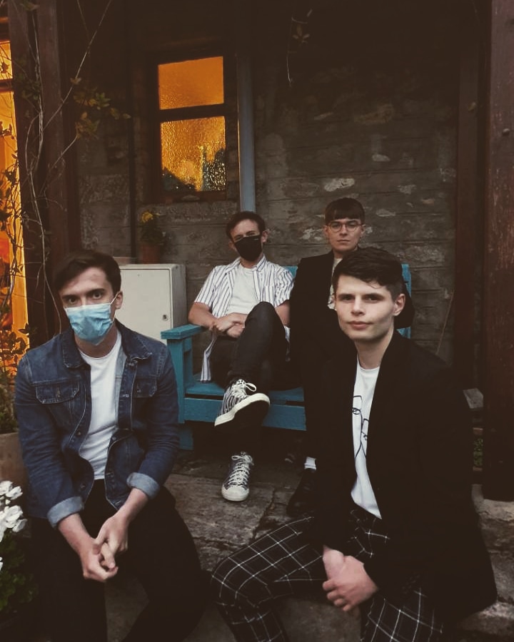Four young men sitting outside a rustic building, two wearing face masks. One man is in a denim jacket, another in a black blazer, with a blue chair and a window in the background.