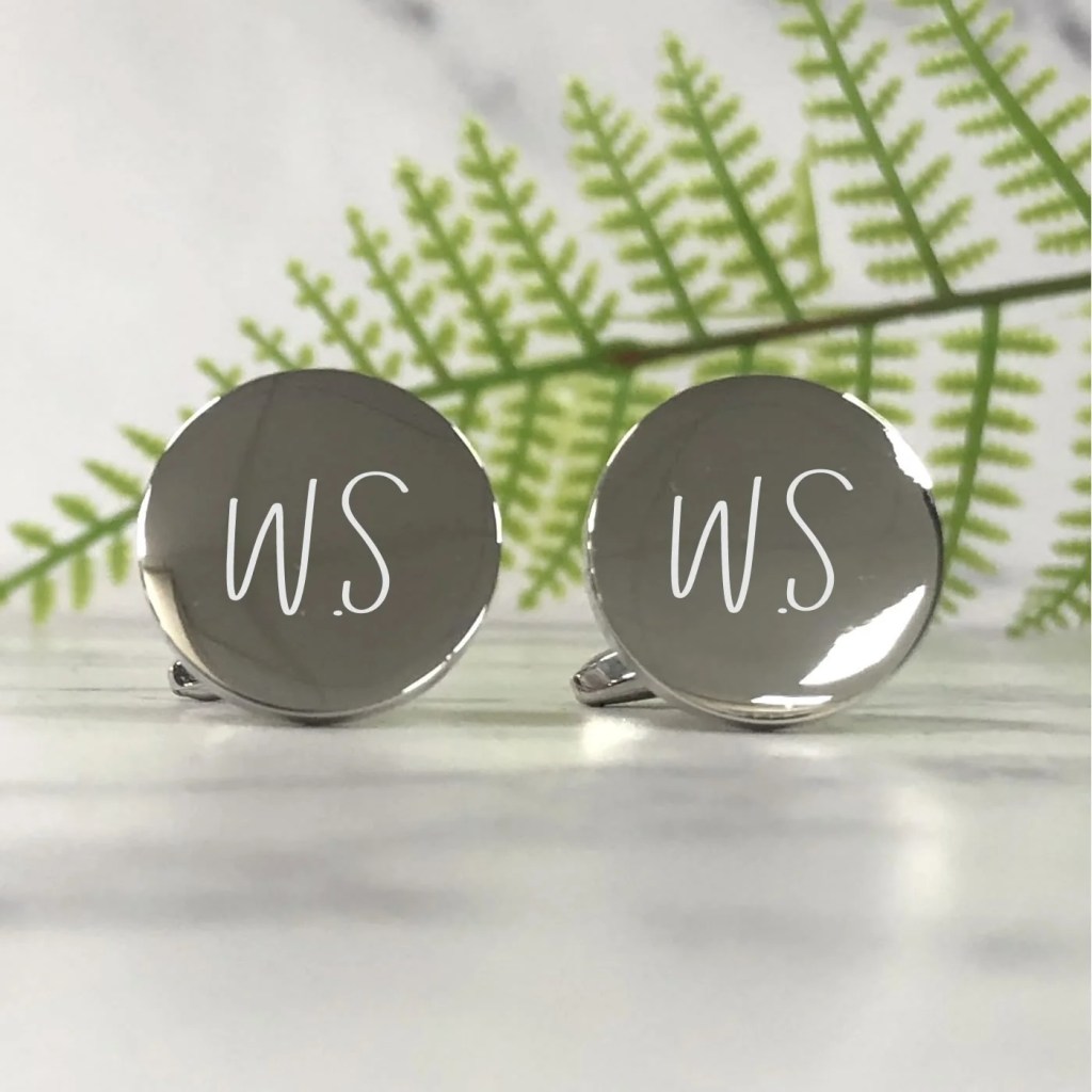 Two circular silver cufflinks with the initials W and S etched into them in a lighter colour. A green fern leaf can be seem behind them. They are displayed on a light surface.