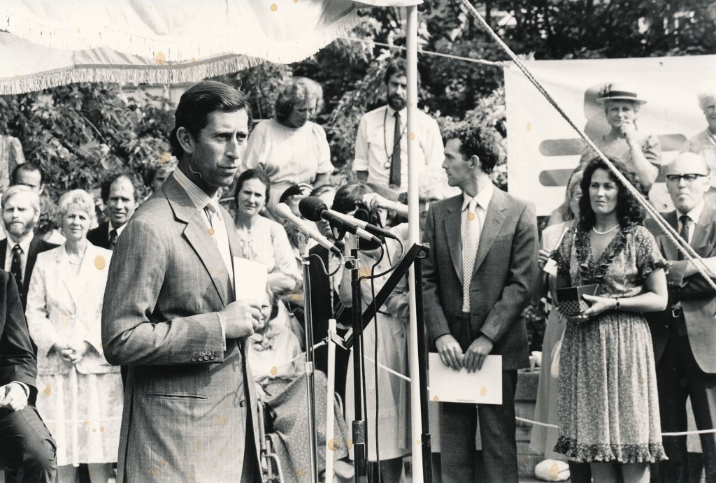 A black and white photograph of a formal speaking event featuring Prince Charles in a suit standing at a microphone, addressing an audience. Several individuals are present behind him, facing the speaker. The setting appears to be outdoors, with a decorative canopy overhead.