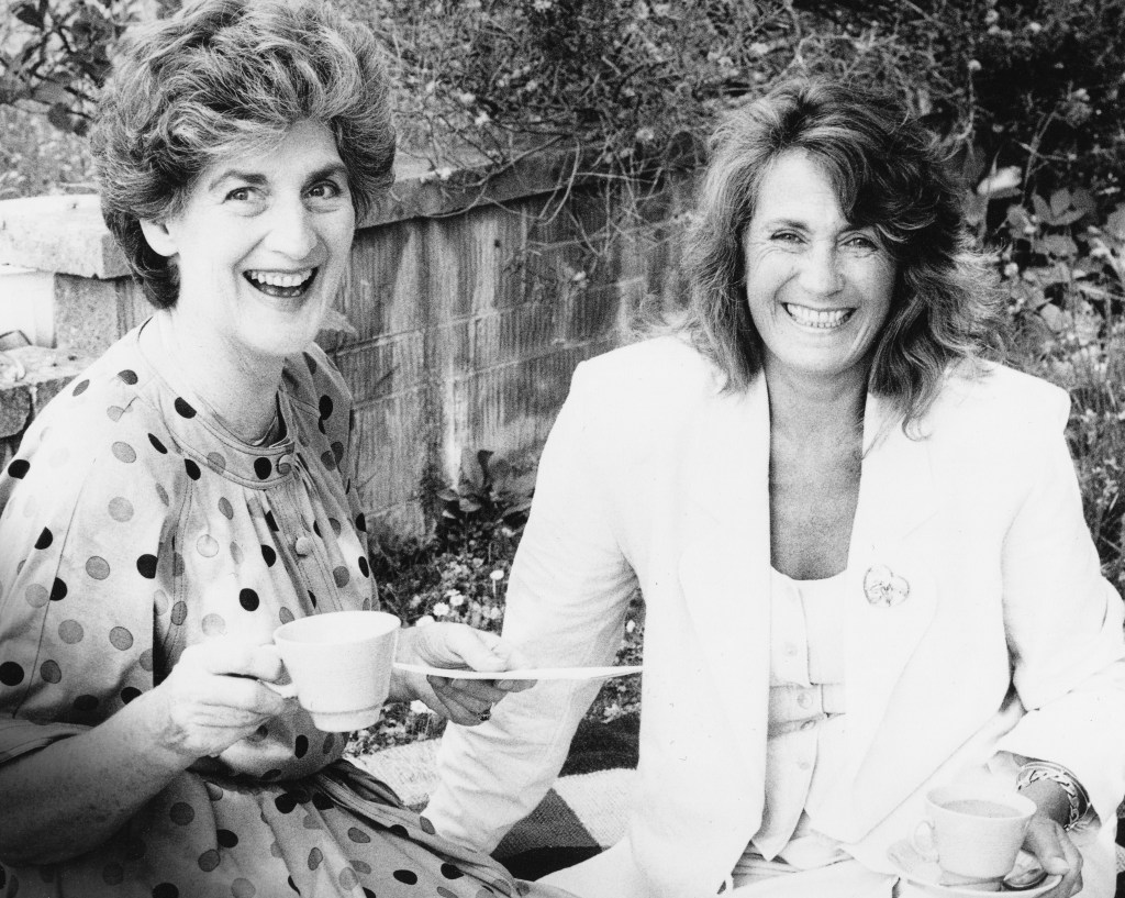 A black and white photo of two smiling women sitting outdoors, enjoying tea and each other's company, dressed in casual outfits, with one wearing a polka dot dress and the other in a white blazer.