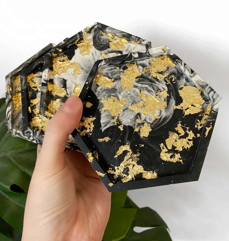 A hand holding four hexagonal coasters featuring a black and white marbled design with gold leaf accents.