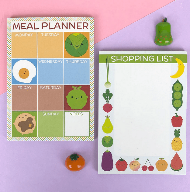 A colourful meal planner and shopping list pad, featuring cute food illustrations, arranged on a pastel purple background. The meal planner is designed with sections for each day of the week, while the shopping list features borders of various fruits and vegetables.