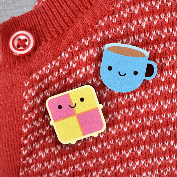 A cute blue coffee mug and a yellow and pink square Battenberg cake icon with a smiling face, pinned to a red and white knitted sweater.