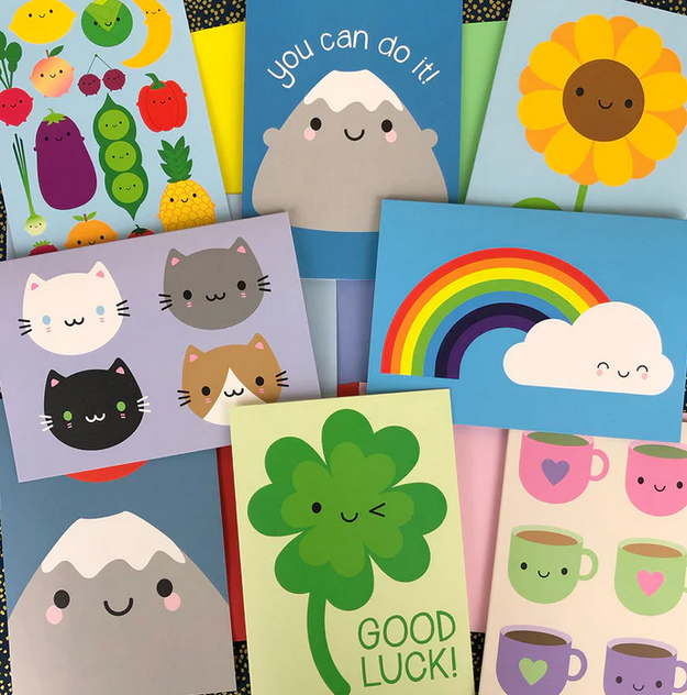 A collection of colourful greeting cards featuring cute illustrations, including smiling fruits, cats, a rainbow, a flower, a mountain, and a shamrock with the phrase 'GOOD LUCK!'