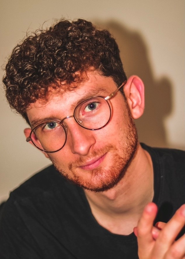 A young man with curly hair and glasses and a black t-shirt is looking directly at the camera with his hands close together.