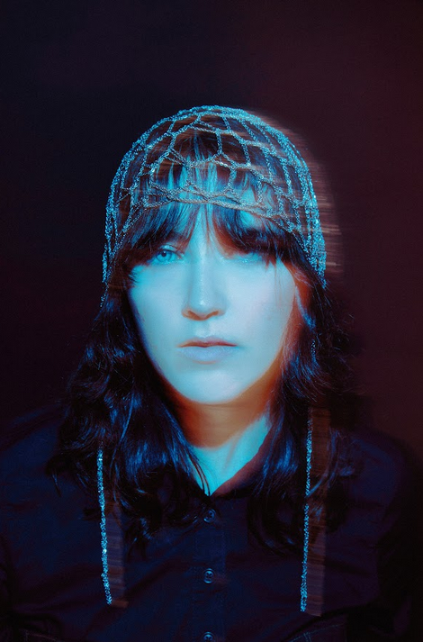 A woman with long dark hair wearing a decorative headpiece, gazing directly at the camera against a dark background with a blue tint.