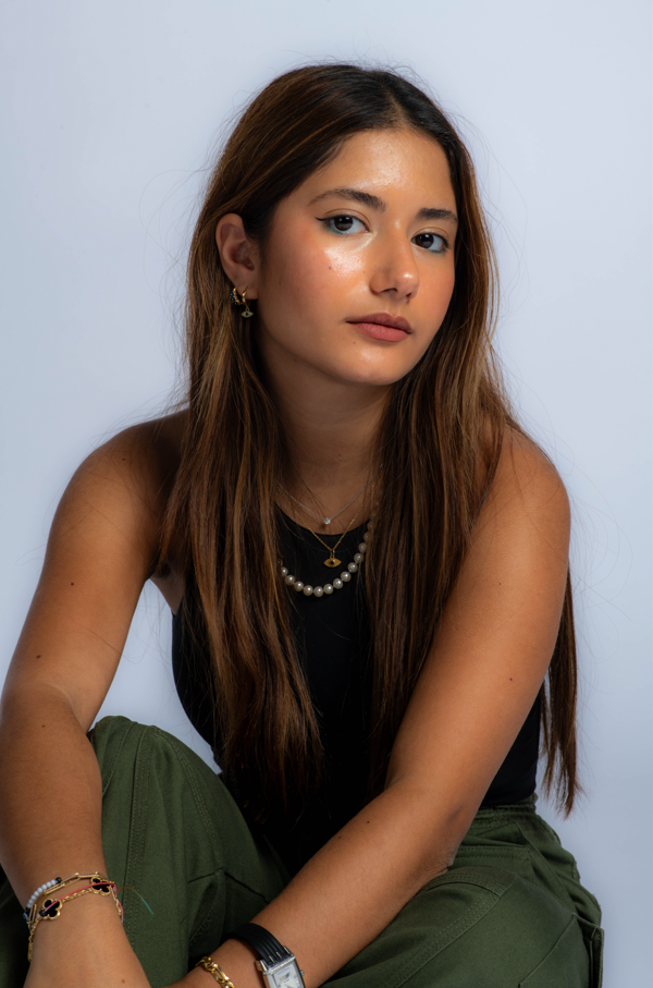A young woman with long brown hair, wearing a black tank top and green trousers, sitting with her arms resting on her knees against a plain white background. She has a natural makeup look and accessorises with multiple necklaces and bracelets.