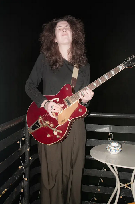 A woman playing a red electric guitar on a balcony at night, showing a joyful expression while surrounded by decorative string lights.