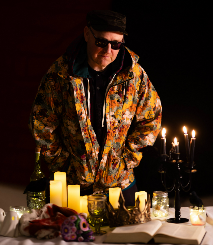 A man wearing a colourful floral jacket and sunglasses, standing behind a table adorned with candles, a bottle, glassware, and a crown, set against a dark background.