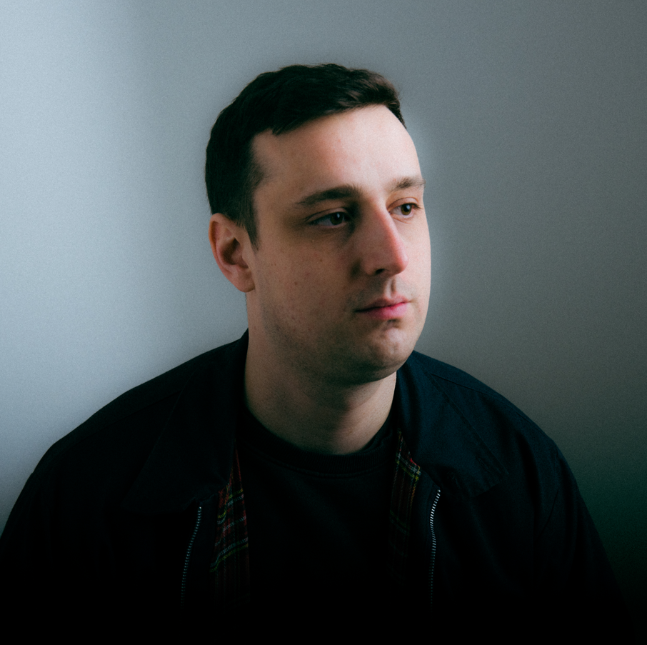 A young man with short dark hair, wearing a dark jacket and dark t-shirt, sits against a light grey wall, gazing thoughtfully to the side.
