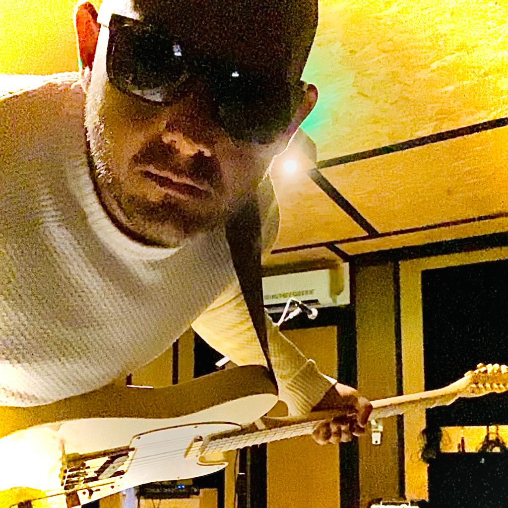 A man wearing sunglasses poses with an electric guitar in a music studio setting.