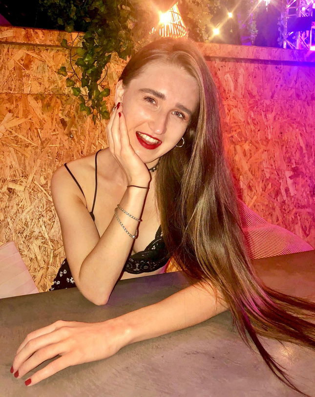 A young woman with long hair and red lipstick resting her face on her hand, smiling at the camera, in a dimly lit setting with colourful lights and wooden background.