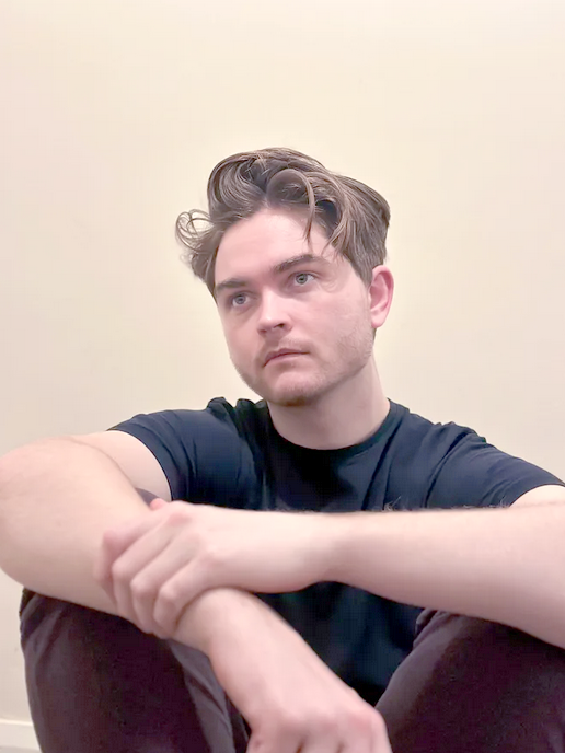 A young man sitting on the floor with his knees drawn up, looking thoughtfully to the side. He has short, styled hair and is wearing a blue t-shirt and maroon trousers.