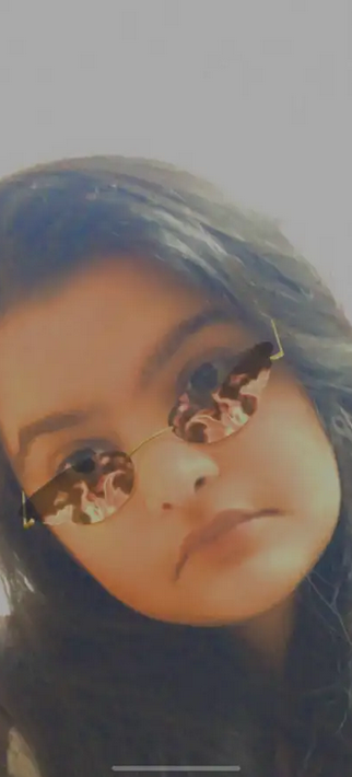 A close-up portrait of a young woman with long dark hair, wearing round sunglasses with a reflective lens. The background is light and blurred.