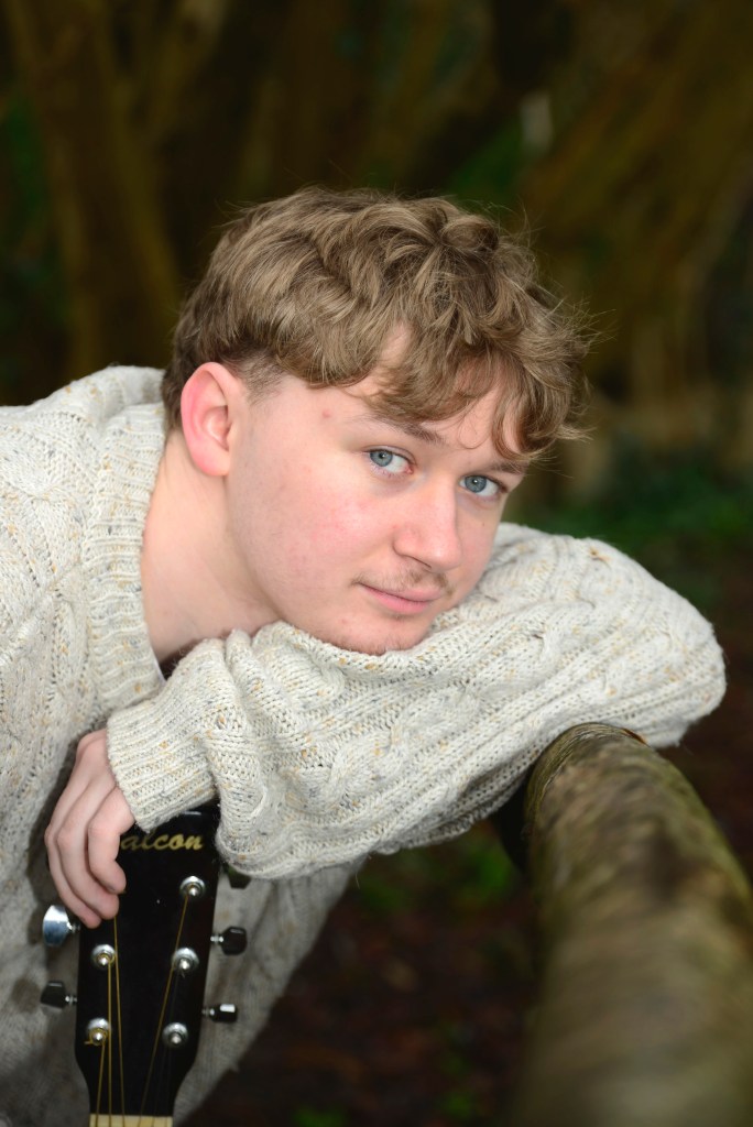 A young man with light curly hair and blue eyes rests his chin on his left arm which rests on a wooden railing, wearing a cream-coloured knitted jumper and holding an acoustic guitar by his side. The background features blurred greenery.
