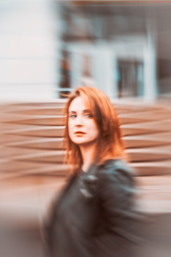 A woman with long red hair wearing a black leather jacket is walking passed a blurred background of stairs and buildings.