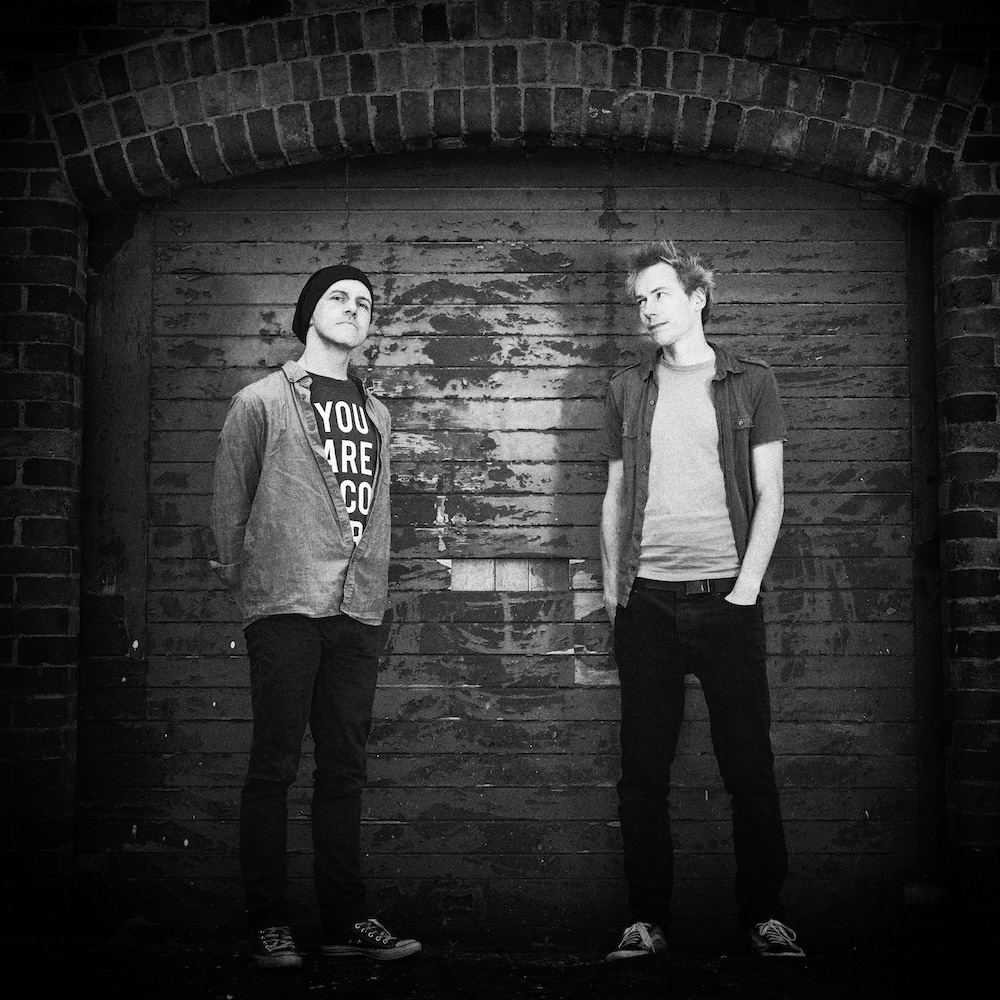 Two young men standing in front of a rustic wooden door with a brick archway, one wearing a beanie and a t-shirt with obscured letters, and the other in a casual shirt, both looking at each other.