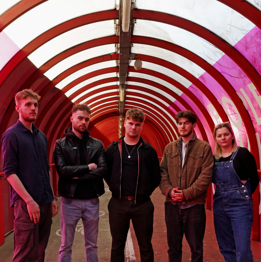 Five individuals standing side by side in a red arched tunnel. The group consists of four men and one woman, with varying hairstyles and outfits. The tunnel has bright red and pink lighting.