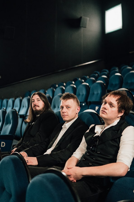 Three men in formal attire seated in an empty theatre, with blue seats in the background.