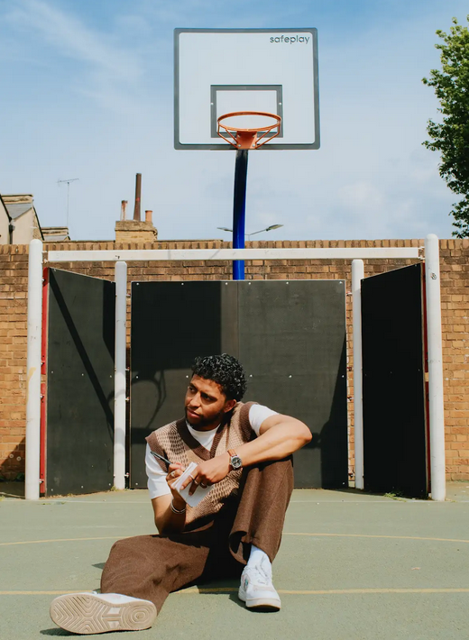 A young man sitting on a basketball court, in front of a hoop, wearing a stylish outfit with a vest and brown trousers, holding a pen and notepad, looking thoughtfully away from the camera.